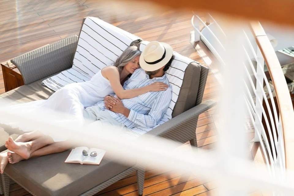 An adult couple relaxes on a sunbed on a wood deck with a book and sunglasses, enjoying leisure time outdoors on a sunny day.