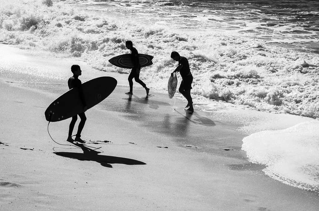 Three surfers walk along a sandy beach toward ocean waves, carrying surfboards, in a black and white shot of beach lifestyle.