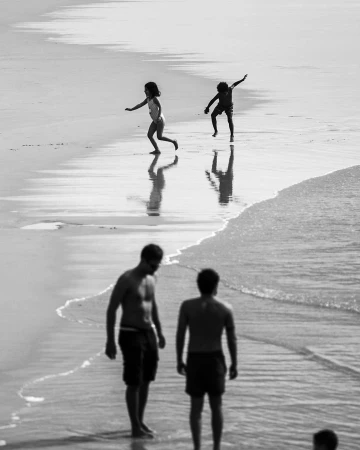 Black and white beach photography by Doug Menuez showing two children playing in the shallows near two adults.
