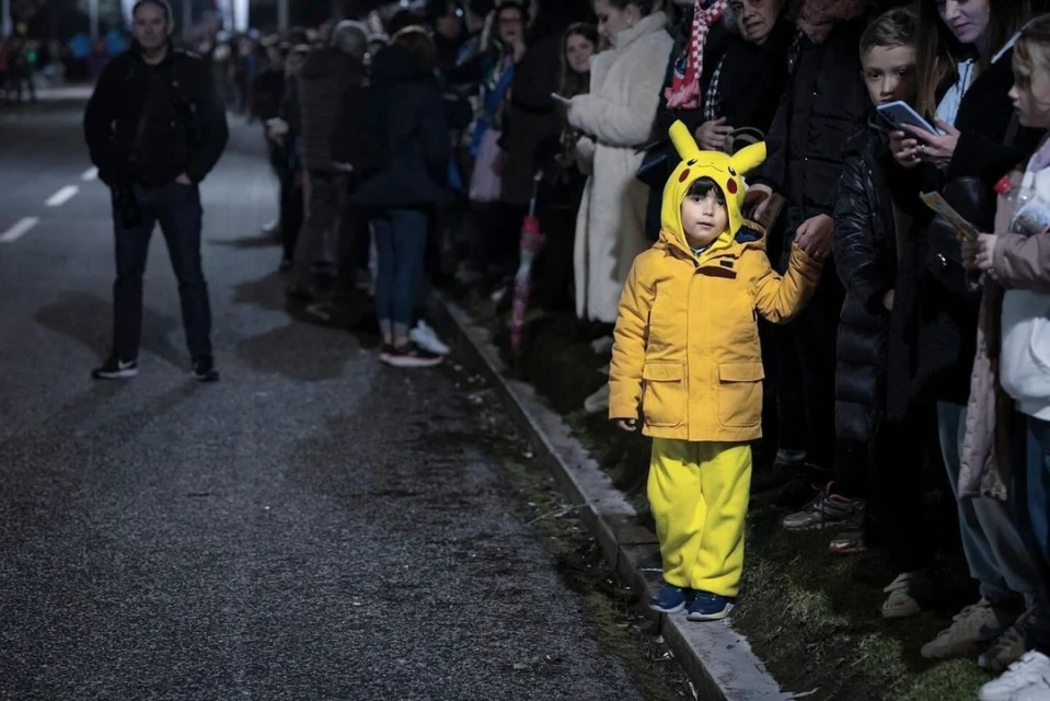 Child in a Pikachu costume stands near a crowd on an urban street at night, wearing a yellow hooded jacket and matching pants.