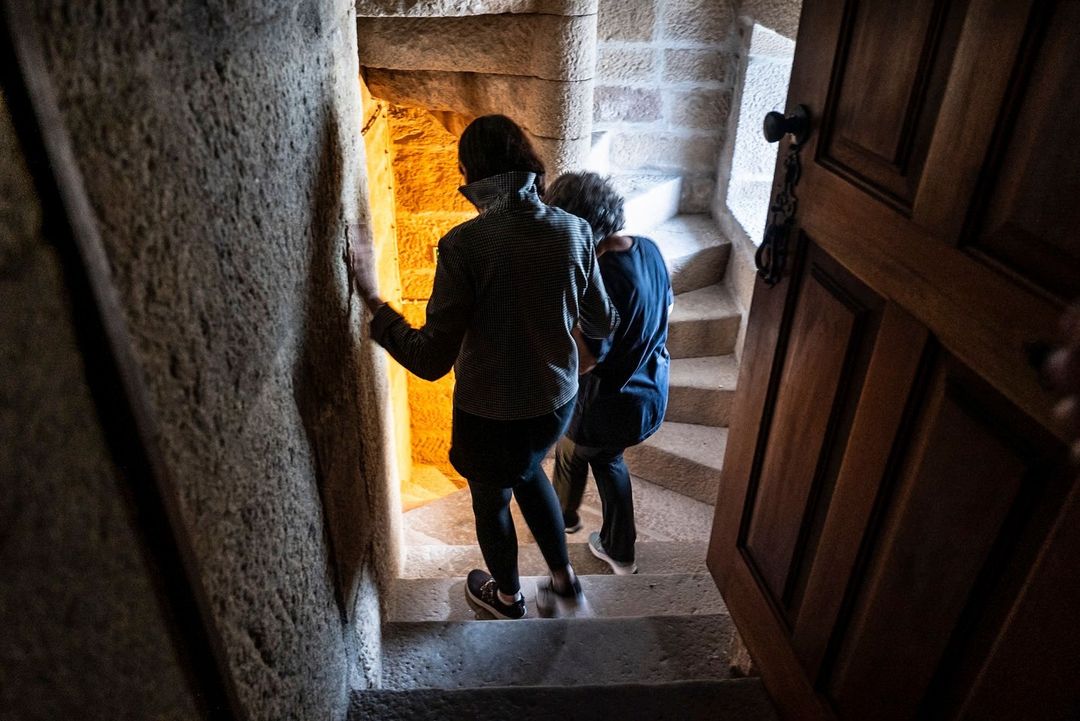 Two people descend a stone spiral staircase inside a building. A partially opened wooden door is visible on the right.