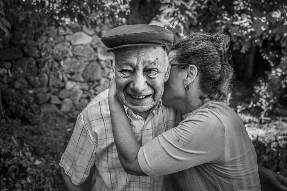 Black-and-white portrait of a smiling elderly man in a cap receiving an affectionate kiss from a woman outdoors, in natural lighting.