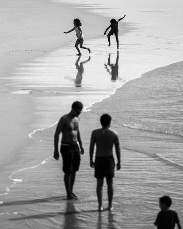 Black and white photograph by Doug Menuez of children running and adults standing on a wet sandy beach.