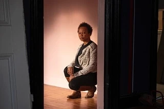 Indoor portrait of a Black woman crouching in doorway with neutral expression, wearing a patterned sweater and dark dress, looking at camera.