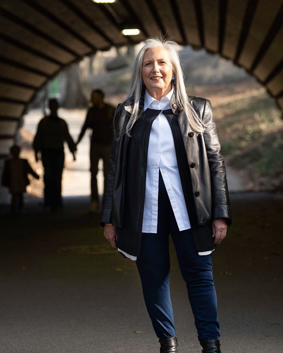 Stylish older woman in a black jacket and white shirt stands in an arched tunnel with a family walking in the background.