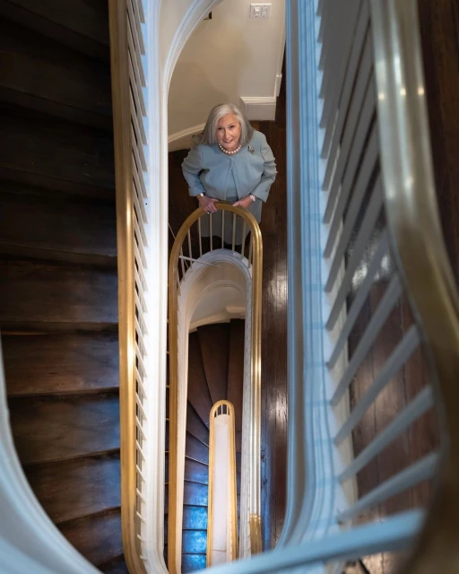 High-angle view of an older woman in a light blue suit standing on a wooden staircase with white railings, looking up at the camera.