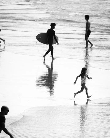 Black and white photograph of surfers and a child running along a wet sandy beach by Doug Menuez