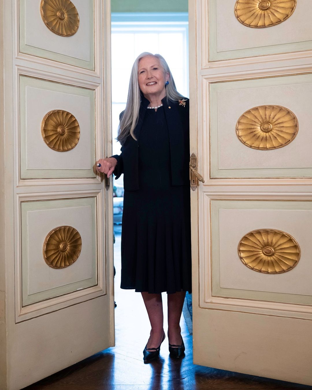 Portrait of a smiling, mature woman with long grey hair wearing a black dress and standing in the doorway of a cream-colored room.