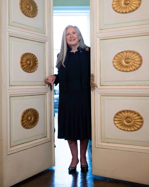 Portrait of a smiling, mature woman with long grey hair wearing a black dress and standing in the doorway of a cream-colored room.