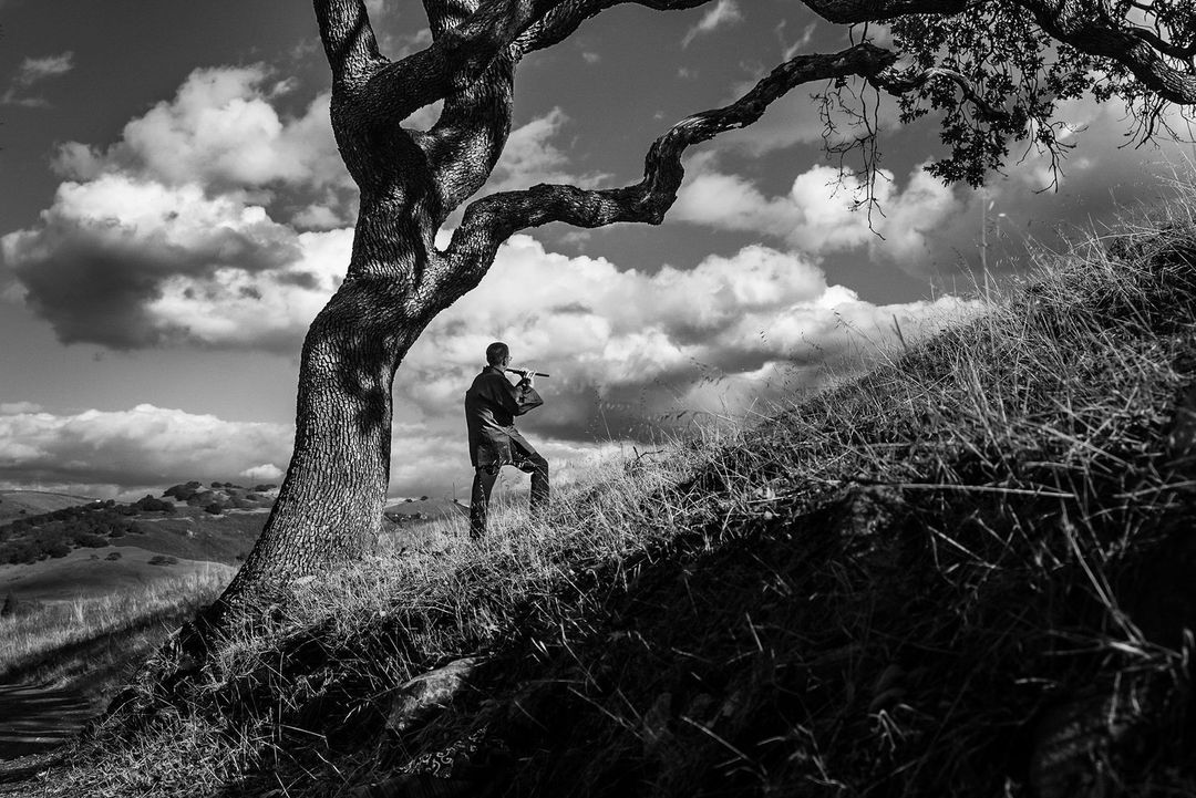 Monochrome outdoor shot of a man playing a flute under a large tree, against a cloudy sky, on a grassy hillside.