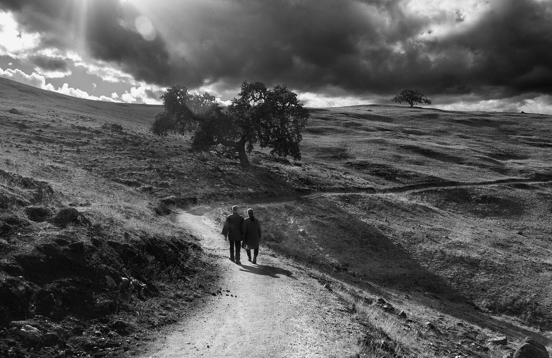 Black and white landscape photograph of a couple walking on a dirt path through grassy hills under a cloudy sky with trees.