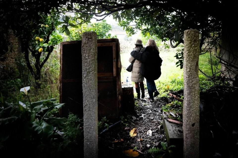 Two women walking on a path in an outdoor garden. The path is lined with stone pillars, greenery, and a wooden structure.