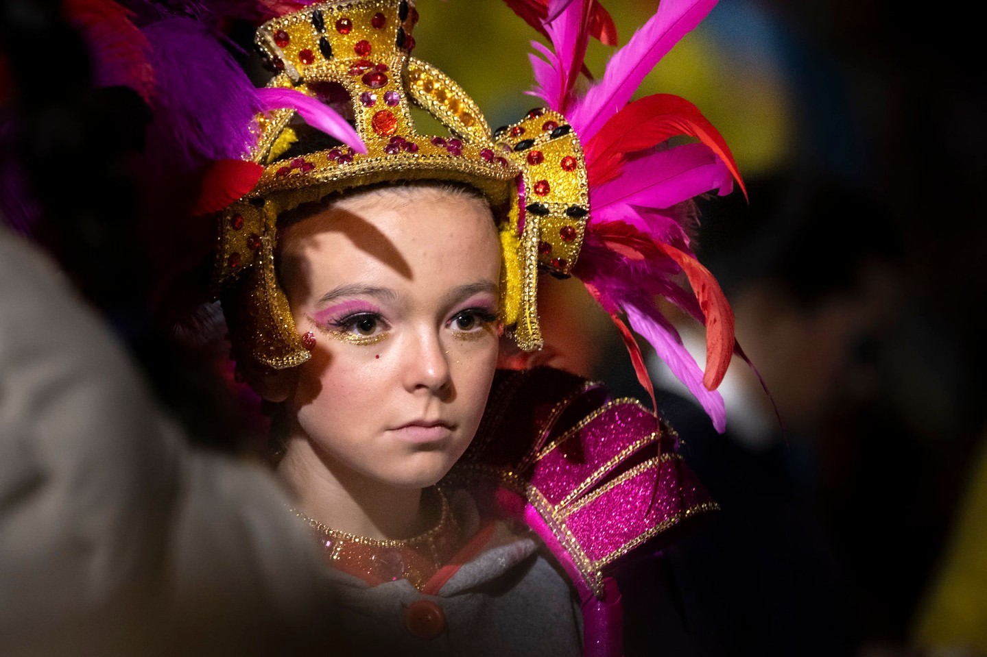 Young girl with glitter eye makeup wearing colorful feathered headdress and shiny necklace, shoulder armor and grey hooded sweater in dim lighting.