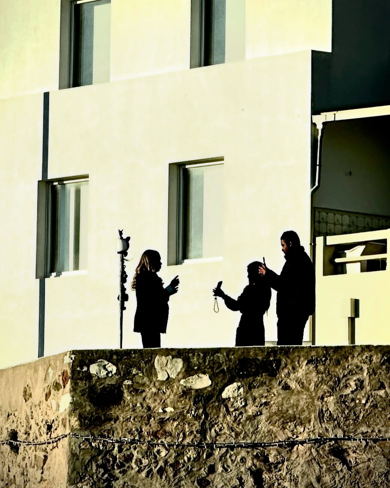 Three silhouetted people on a stone wall against a bright white building, one taking a photo with a smartphone, outdoors.