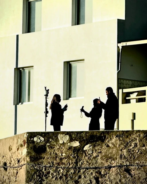Three silhouetted people on a stone wall against a bright white building, one taking a photo with a smartphone, outdoors.