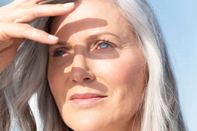 Close-up portrait of an elderly white woman with grey hair shading her eyes from the sun with her hand against a clear blue sky