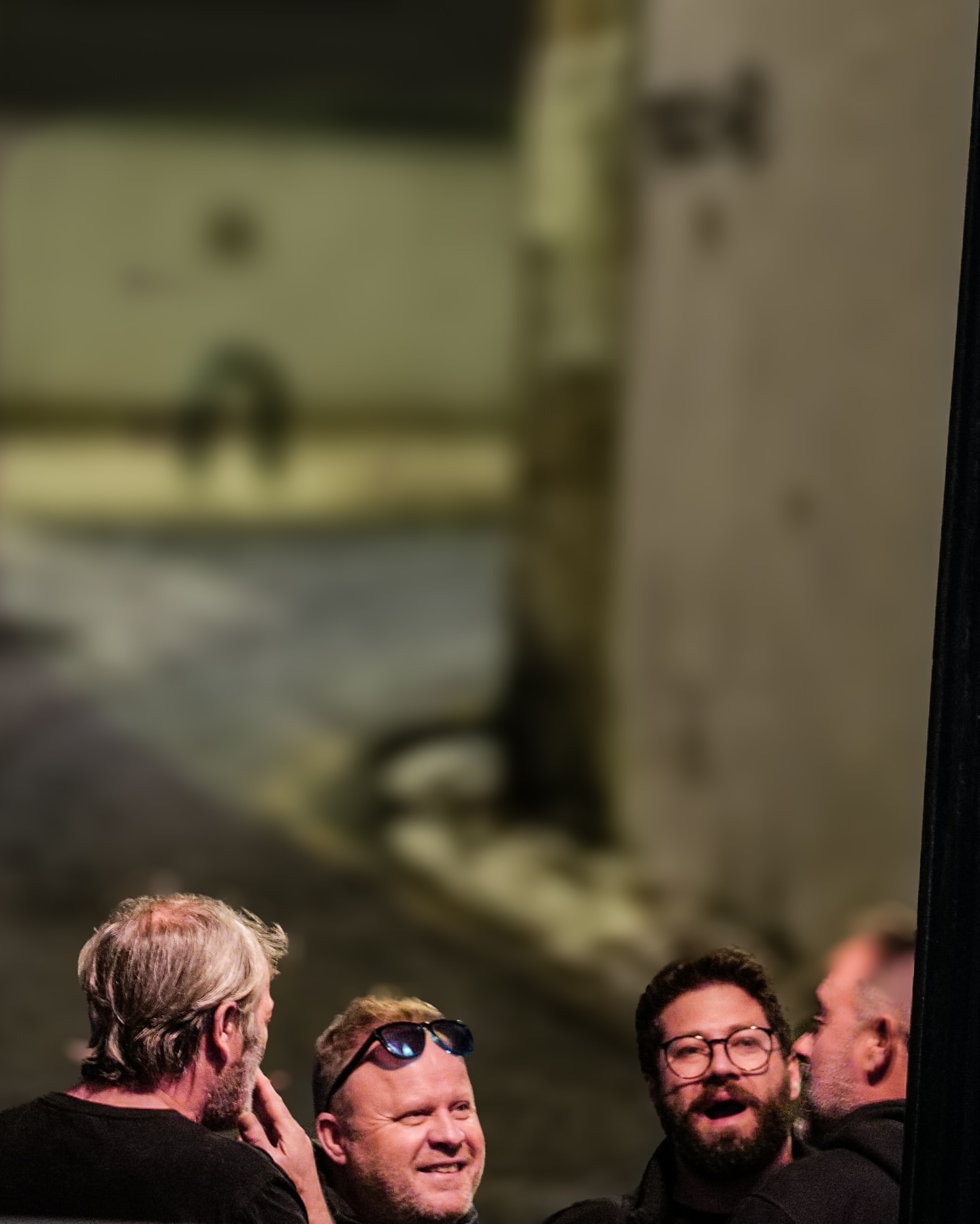 Four adult males gather outdoors in a blurred alleyway, two with glasses, creating a candid moment in a casual setting.