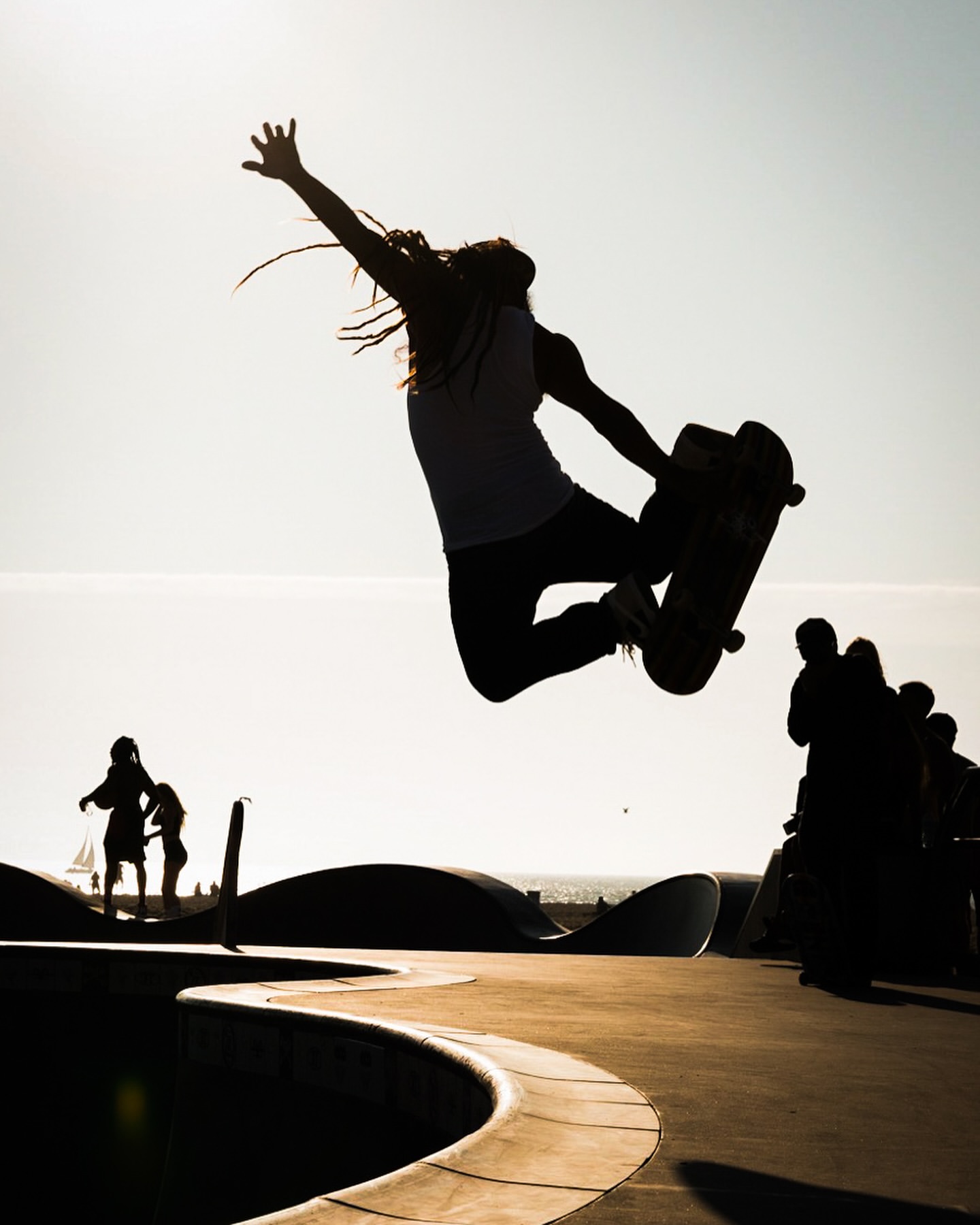 Silhouette of a skateboarder catching air at a skatepark near the beach with silhouetted spectators in the background under a bright sky.