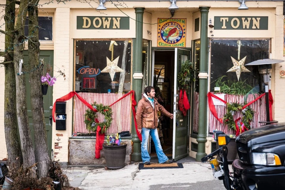 Man in a leather jacket stands in the doorway of Downtown Cafe on a city street, decorated with wreaths and ribbons, car parked nearby.