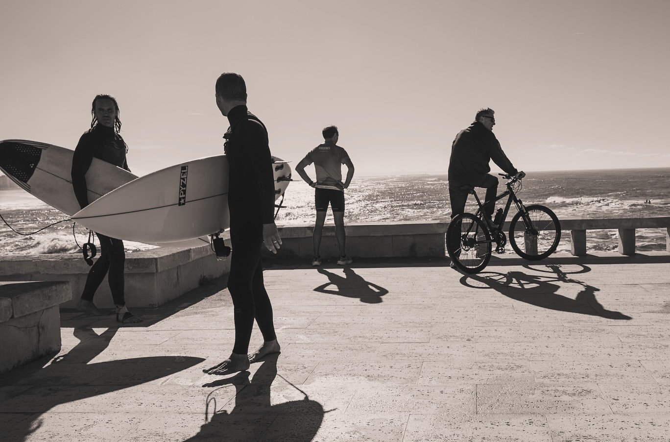 Monochrome shot of two surfers carrying surfboards, a person standing, and another riding a bike by the sea, under a bright sky.