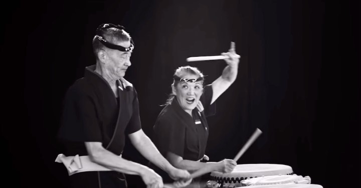 Two musicians play taiko drums enthusiastically in a black and white photo studio setting with a dark background and drumsticks.