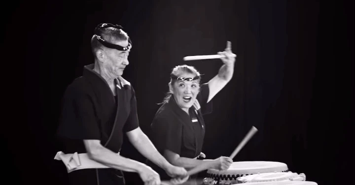 Two musicians play taiko drums enthusiastically in a black and white photo studio setting with a dark background and drumsticks.