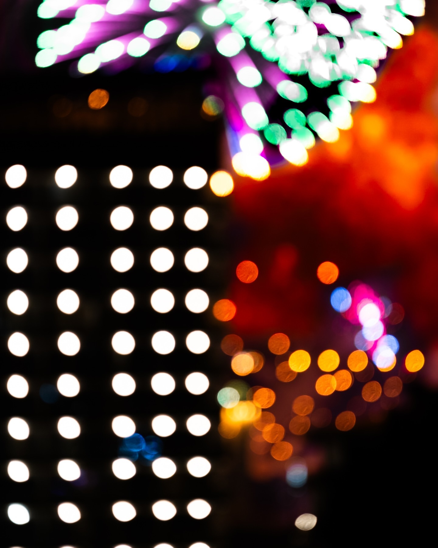 Abstract nighttime display with bright lights in blurred patterns, featuring fireworks and a building facade lit with evenly spaced lights.