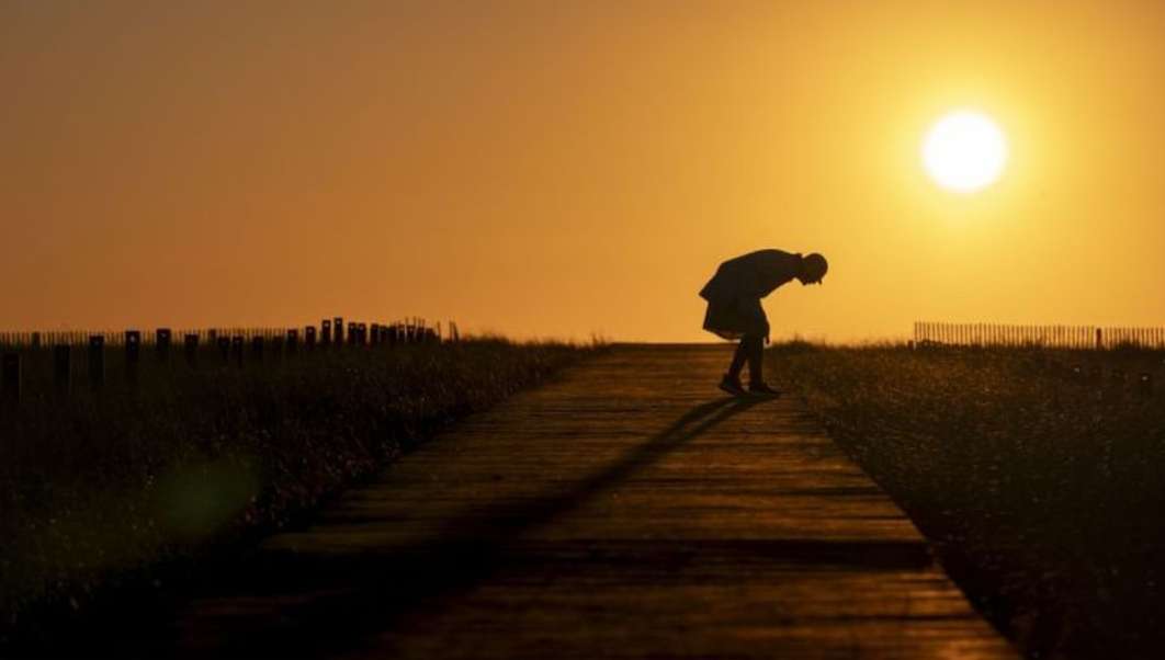 Silhouette of a person bending over on a path with grass on either side, against a bright orange sunset sky.