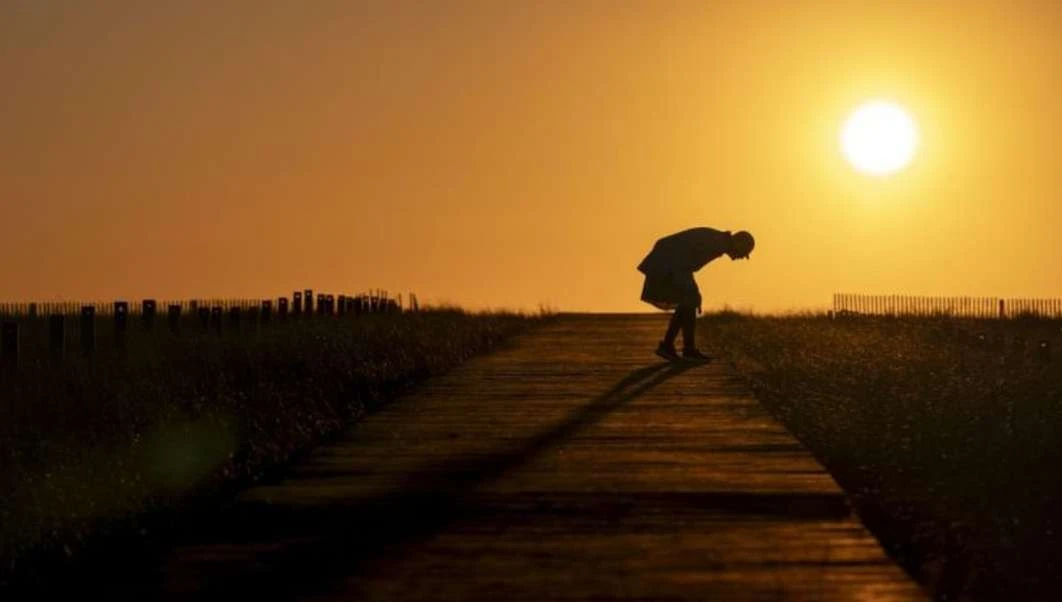 Silhouette of a person bending over on a path with grass on either side, against a bright orange sunset sky.