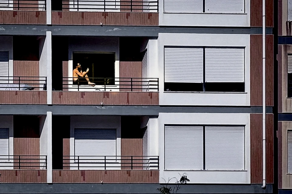 Man sitting on a balcony ledge in an urban apartment complex. Balconies and windows form a symmetrical grid-like facade, day light.