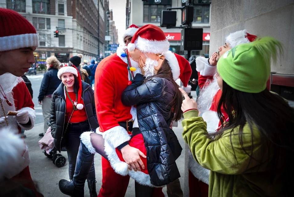 Festive street scene with a group of people in Santa Claus costumes and a woman in black getting a kiss in an urban setting.