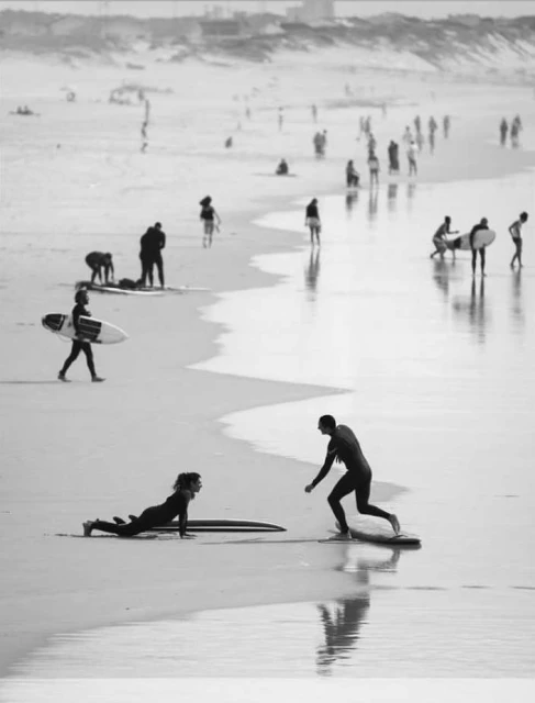 Monochrome shot of surfers on a sandy beach, two in wetsuits, with many people in the distance, reflected in the wet sand.