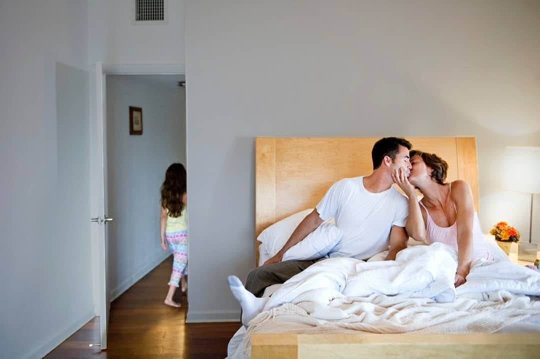 Man and woman kissing on a bed while a child enters the bedroom through an open doorway in the background