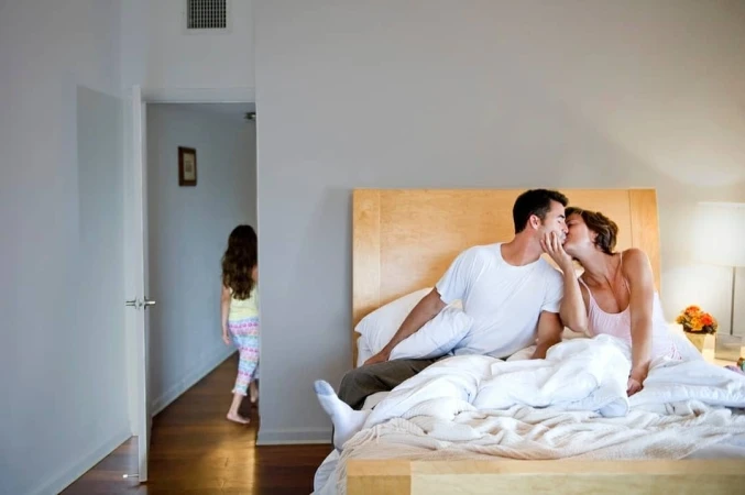 Man and woman kissing on a bed while a child enters the bedroom through an open doorway in the background