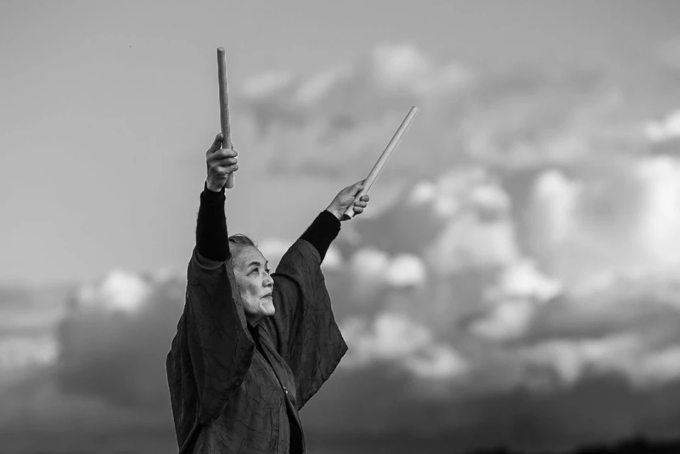 Elderly woman stands outdoors raising drumsticks against a cloudy sky in a monochrome photograph, expressing tradition and strength.