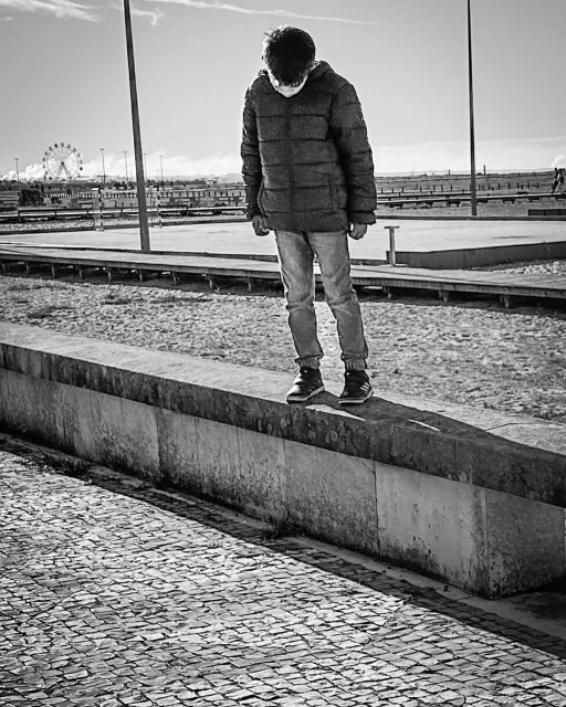 Teenager standing on a concrete ledge in a black and white outdoor shot, with a distant ferris wheel and partly cloudy sky.