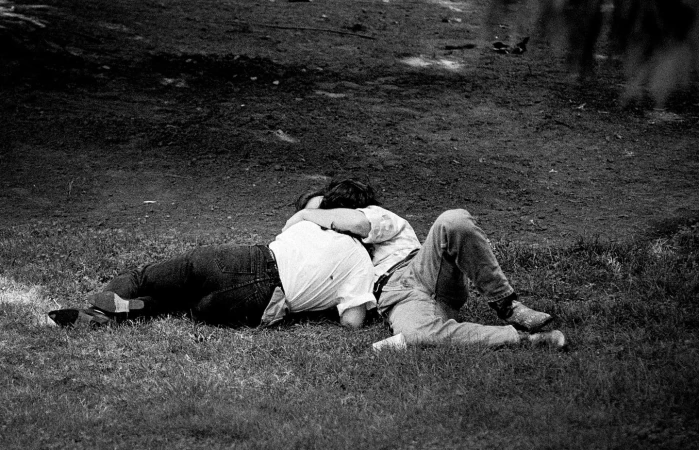 Black and white photograph of two people embracing and lying together on a grassy lawn outdoors.