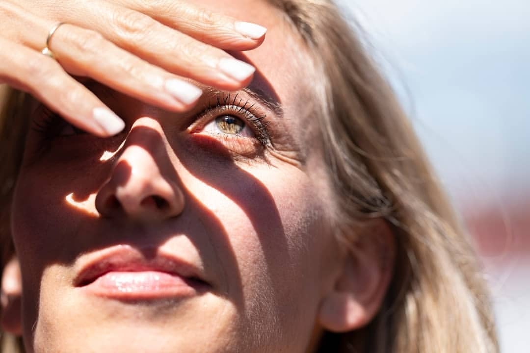 Close-up of a woman shielding her face from the sun with her hand. The sunlight creates shadows across her face and highlights her eye.