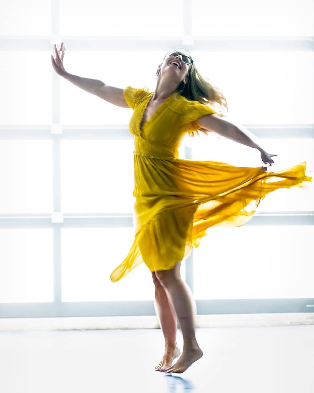 Woman in a flowing yellow dress spinning joyfully with arms outstretched against a bright grid window backdrop