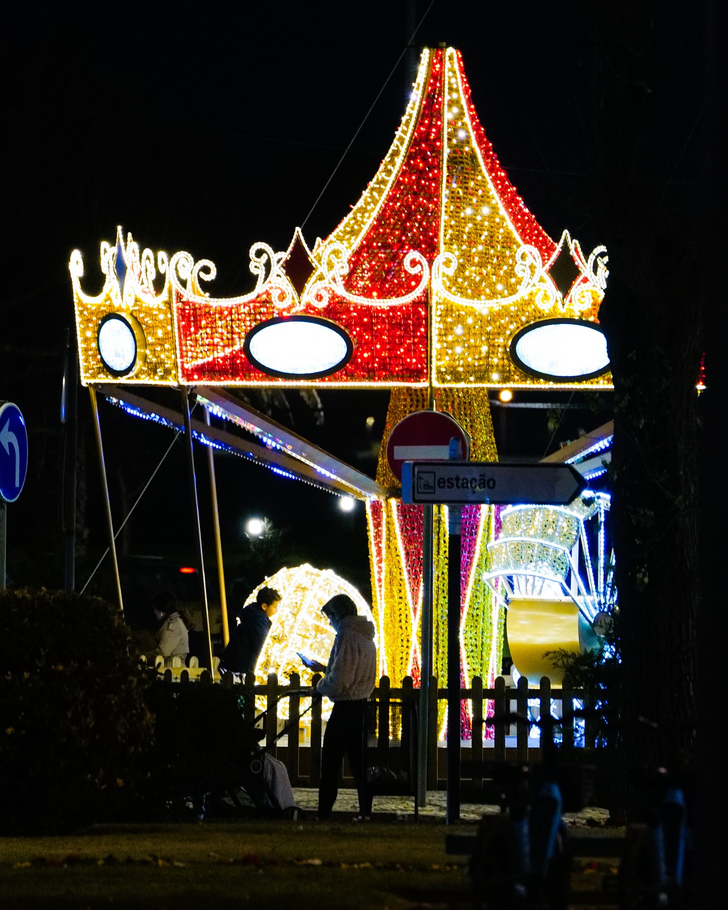 Night scene featuring illuminated carousel-style light display in red and gold, with two people standing in front of it.