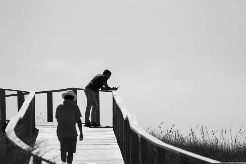 Two adult males on an elevated wooden walkway bordered by wooden railings with tall grass in a black and white outdoor setting.