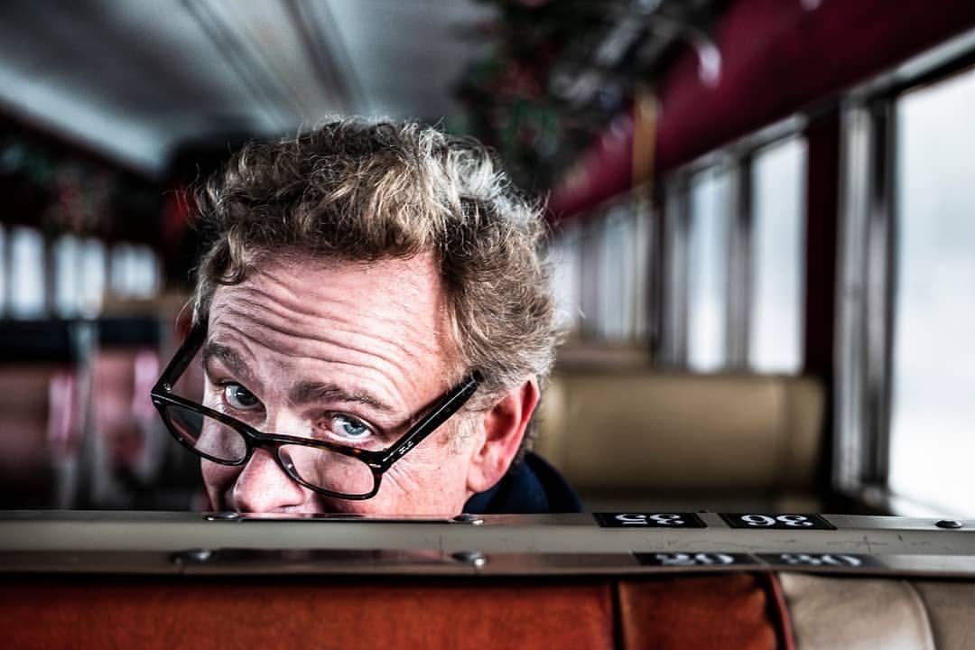 Close-up portrait of an adult male with curly blond hair wearing glasses, peering over an orange and beige seat on a train car.