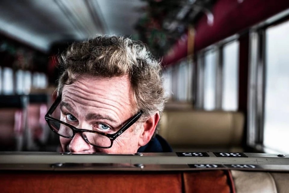 Close-up portrait of an adult male with curly blond hair wearing glasses, peering over an orange and beige seat on a train car.