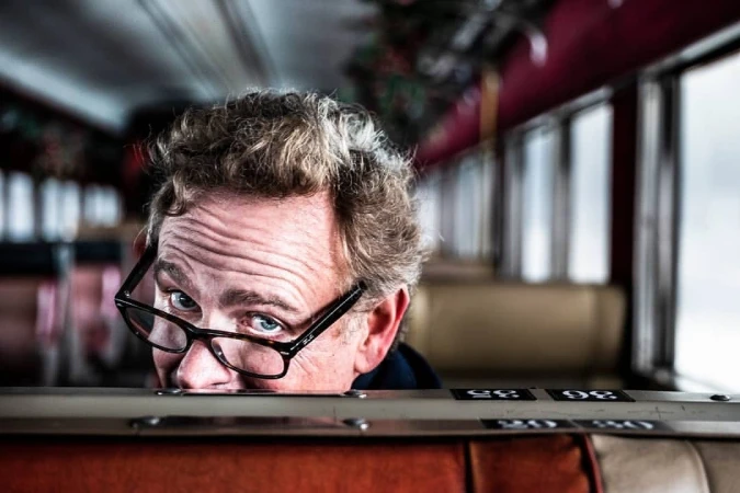 Close-up portrait of an adult male with curly blond hair wearing glasses, peering over an orange and beige seat on a train car.