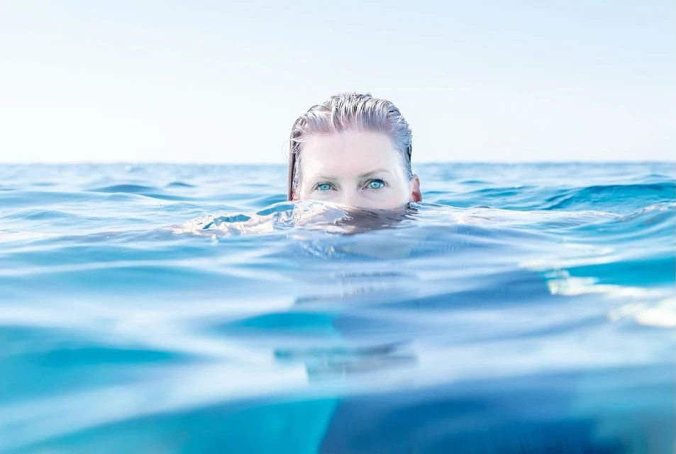 Adult woman with fair skin and blue eyes swims in turquoise water under a clear sky, with her head partially submerged.