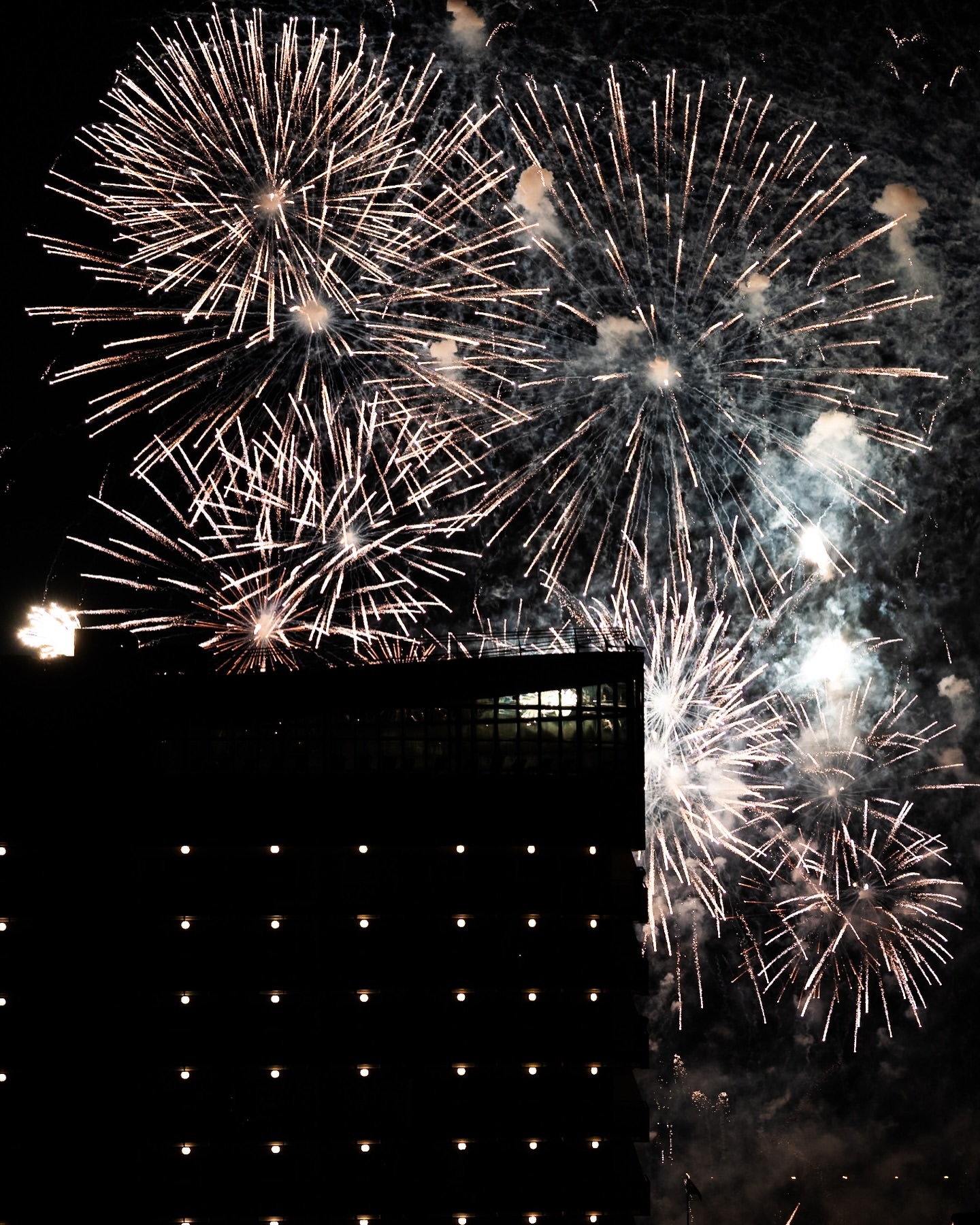 Fireworks exploding in the night sky behind a dark, illuminated building, creating a dynamic and celebratory visual display.