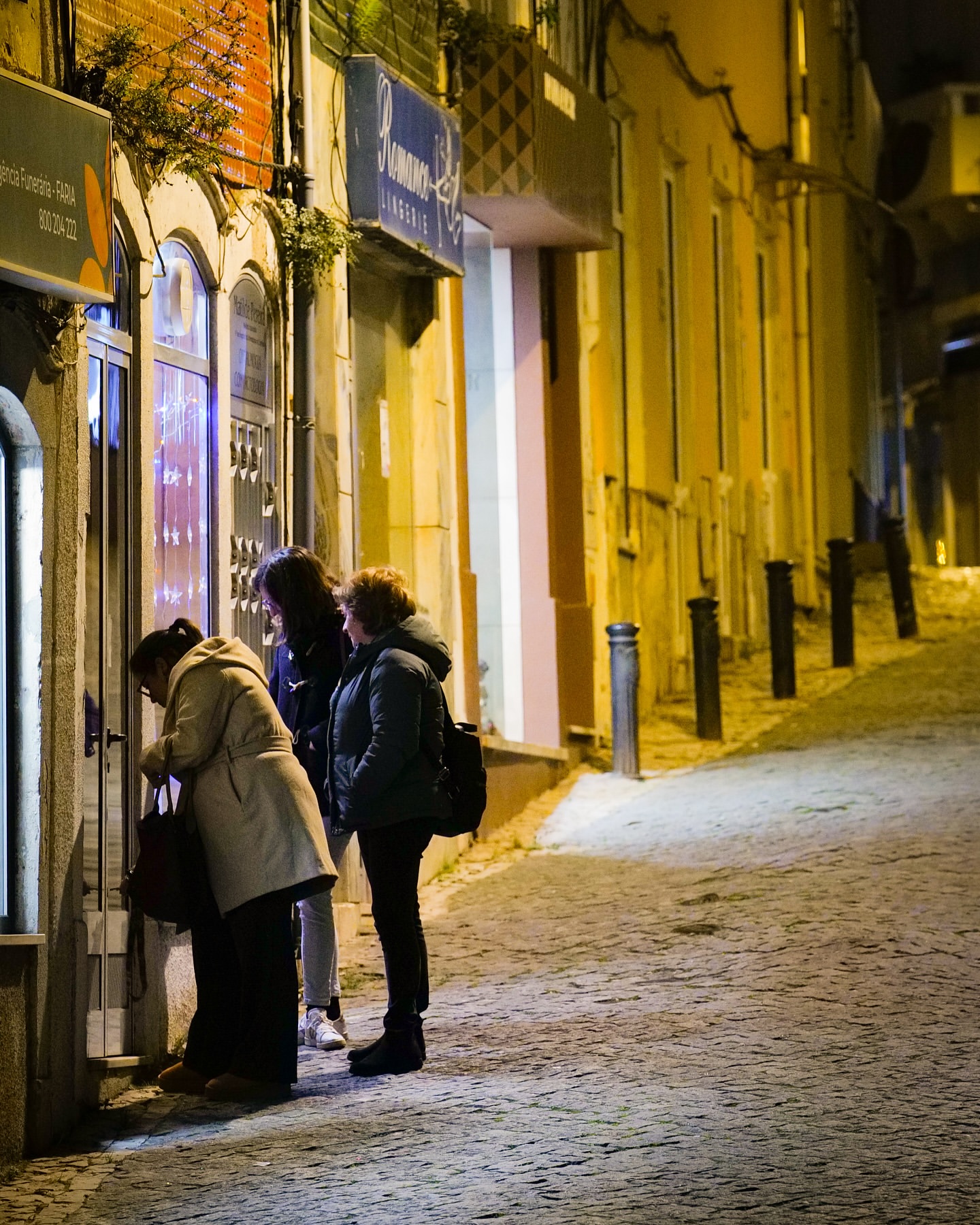 Three women in coats window shopping at night on a cobblestone street with warm lighting illuminating the building facades.