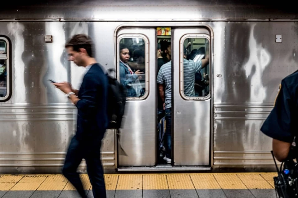 People boarding a metallic subway train, waiting on a yellow-striped platform, with a security guard partially visible.
