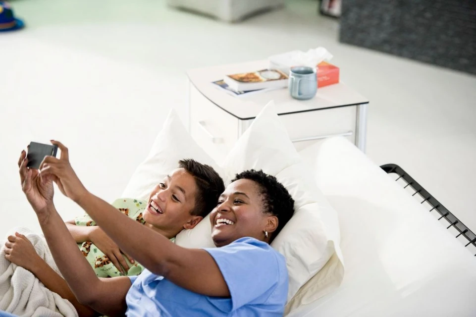 Woman and child take a selfie lying on a white bed. The smiling people are indoors, in soft light, using a cell phone to capture the moment.