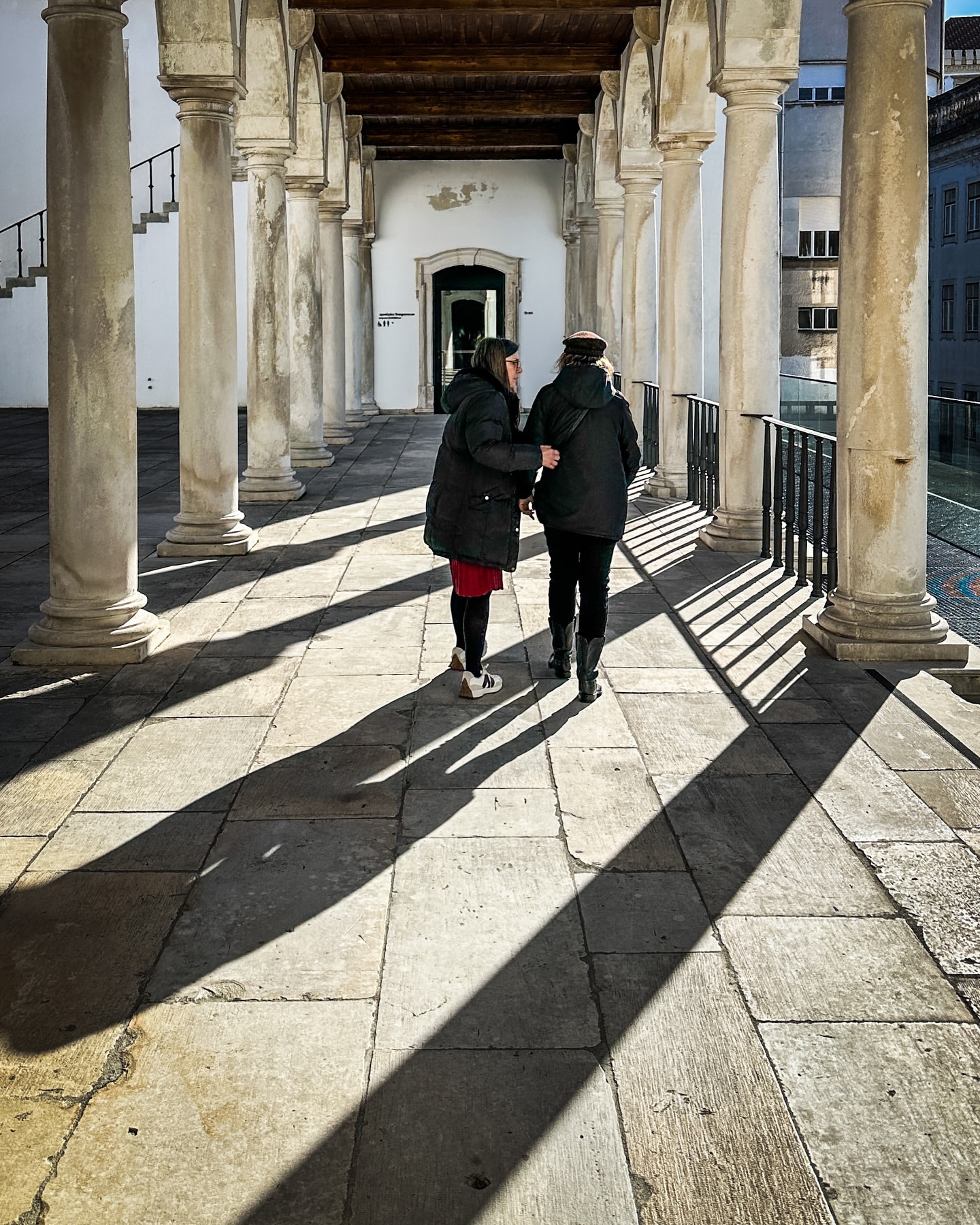 Two adult females walking together through a stone colonnade on a sunny day, casting long shadows on the stone pathway.